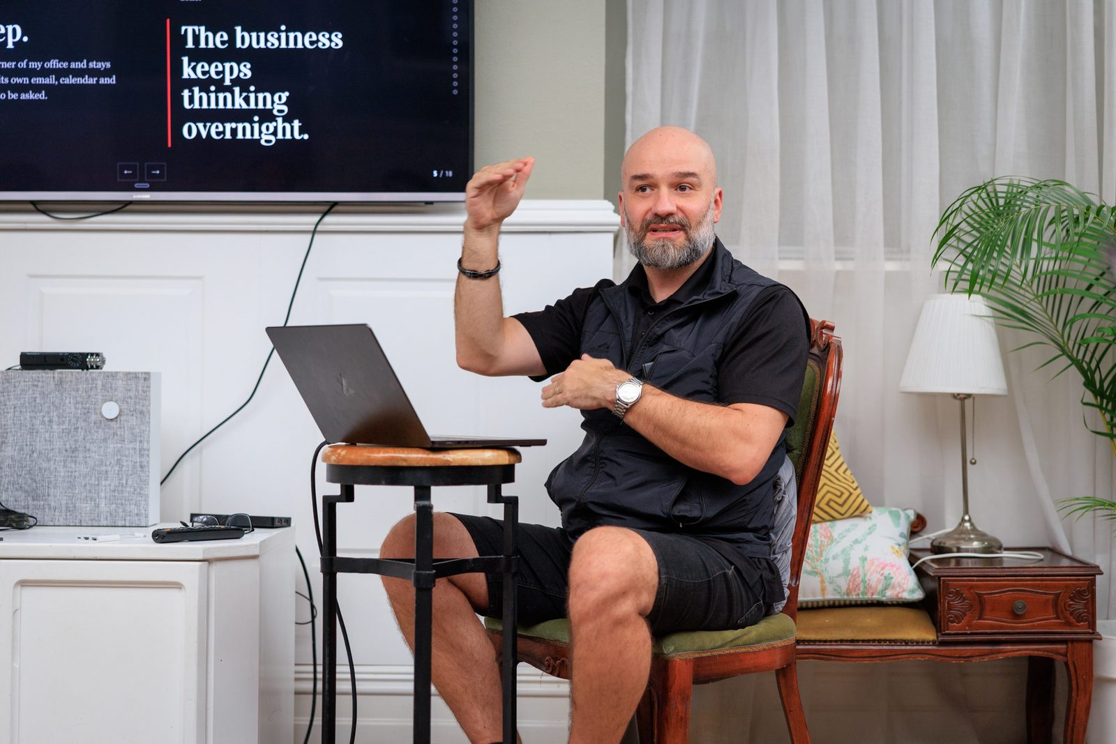 Arek Rainczuk seated with a laptop on a stool, mid-sentence, presenting to the room